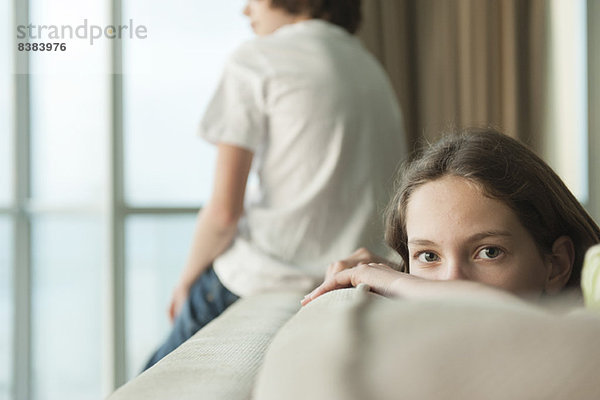 Mädchen auf Sofa sitzend mit Kopf auf dem Arm  Bruder mit Blick aus dem Fenster im Hintergrund