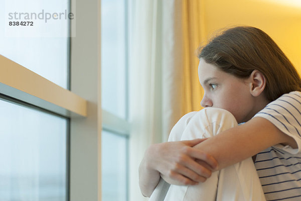 Preteen Mädchen Knie umarmend  aus dem Fenster schauend