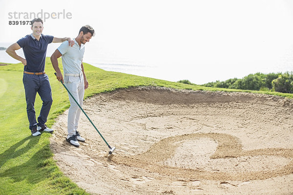 Männer  die in einer Sandfalle auf dem Golfplatz herzförmig rechen.