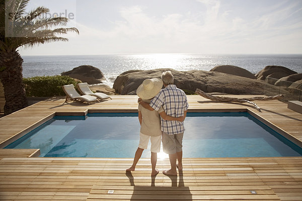 Seniorenpaar umarmt vom modernen Pool mit Blick aufs Meer