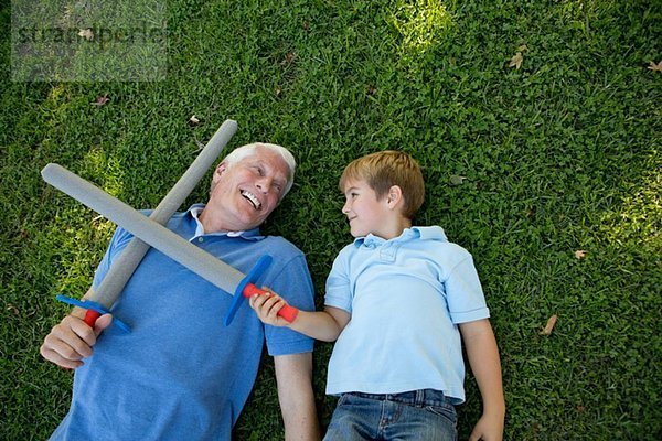 Großvater und Enkel auf Gras liegend mit Spielzeugschwertern