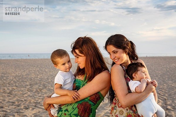 Mütter stehen am Strand und halten Baby und Kleinkind.