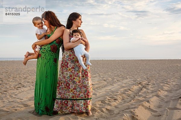 Mütter stehen am Strand und halten Baby und Kleinkind.