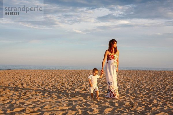 Mutter und Kleinkind gehen am Strand entlang und halten Händchen.