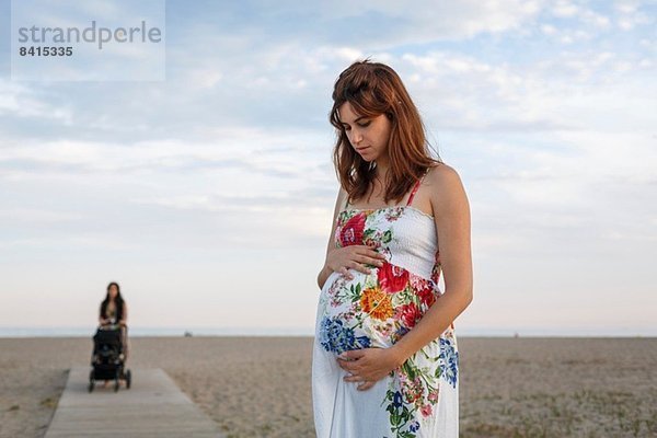 Schwangere Frau steht auf der Strandpromenade  Frau schiebt Kinderwagen im Hintergrund
