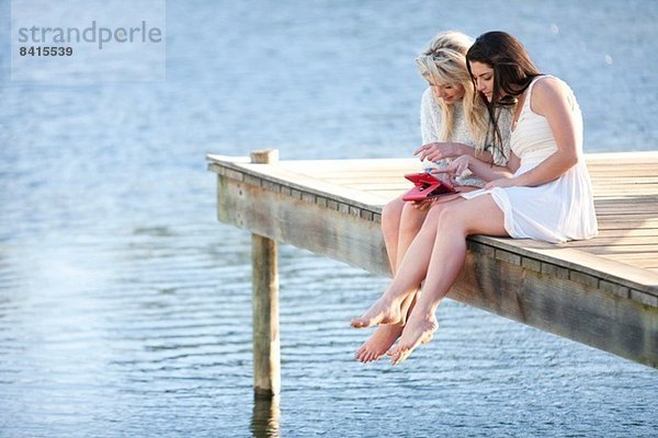 Zwei junge Frauen sitzen auf dem Pier und schauen auf ein digitales Tablett.