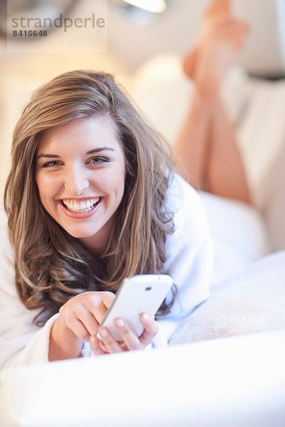 Porträt einer jungen Frau auf dem Bett liegend mit Handy