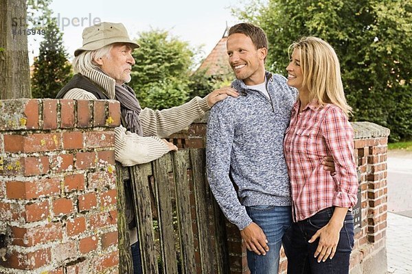 Älterer Mann  der sich auf das Tor stützt und mit einem mittleren erwachsenen Paar redet.