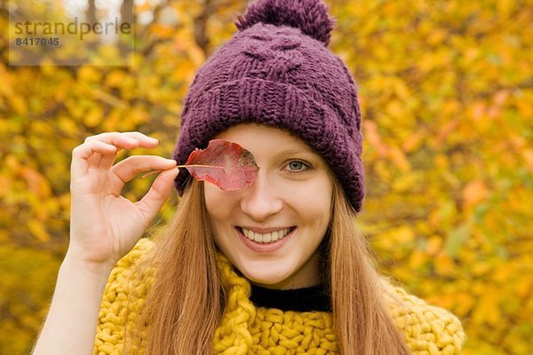 Nahaufnahme einer jungen Frau im Park  die das Herbstblatt über dem Auge hält.