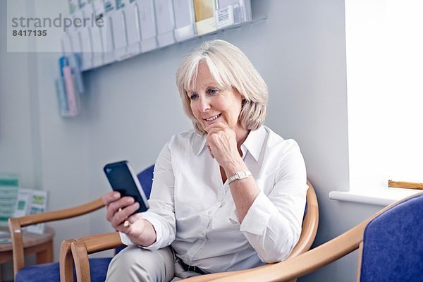 Reife Patientin beim Betrachten des Mobiltelefons im Wartezimmer des Krankenhauses