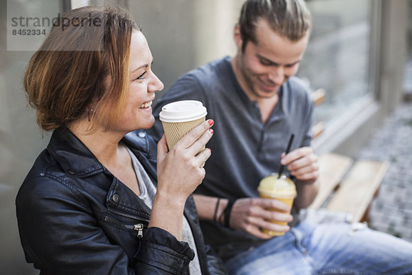 Fröhliche Frau trinkt Kaffee in Einwegbecher mit Mann auf Bank auf dem Bürgersteig