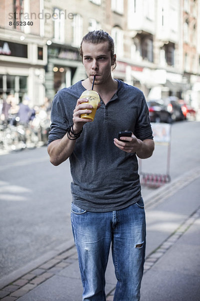 Junger Mann trinkt Saft  während er auf dem Bürgersteig steht.