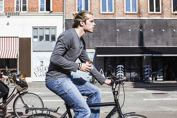 Profilaufnahme eines jungen Mannes  der Burger hält  während er Fahrrad fährt.