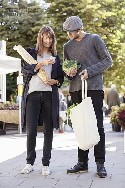 Volle Länge des Paares mit dem Handy beim Einkaufen von Gemüse auf dem Markt