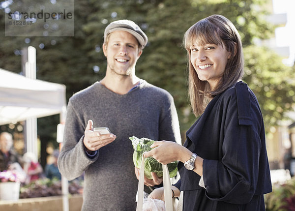 Porträt eines lächelnden Paares mit Handy und Lebensmitteln auf dem Markt