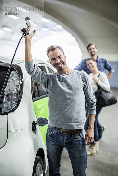 Porträt eines glücklichen Mannes mit elektrischem Ladegerät und Freunden im Hintergrund an der Tankstelle