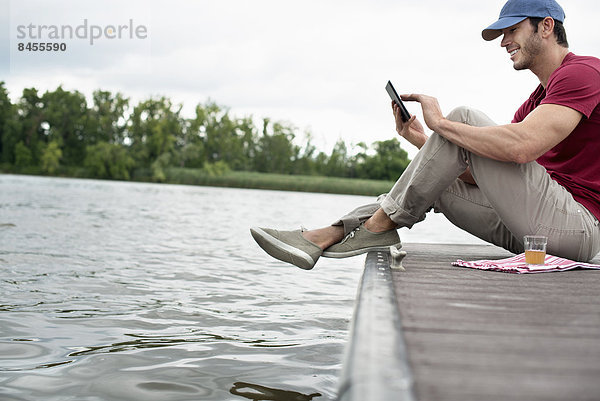 Ein Mann sitzt auf einem Steg an einem See und benutzt ein digitales Tablett.