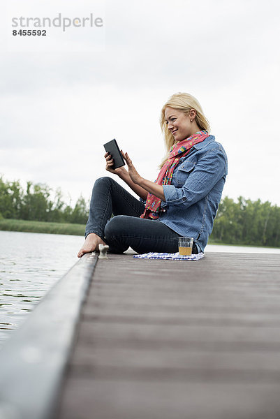Eine Frau sitzt auf einem Steg an einem See und benutzt ein digitales Tablett.