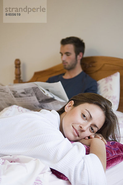 Ein Mann  der im Bett Zeitung liest  und eine Frau  die sich mit einem Lächeln entspannt.