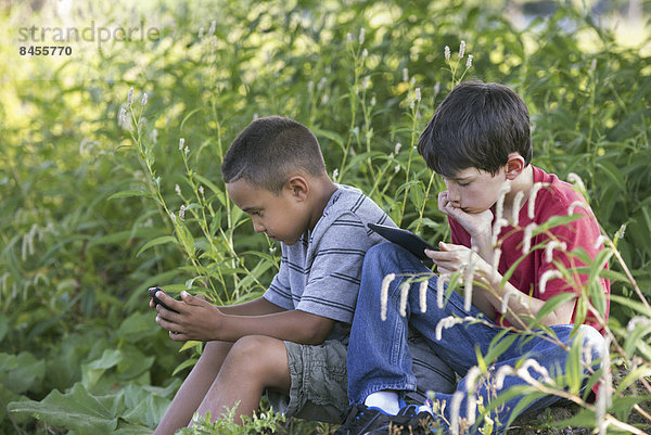 Zwei Jungen sitzen auf einem Feld  einer mit einem Smartphone und einer mit einem digitalen Tablet.