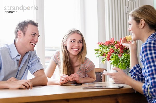 Drei junge Leute sitzen an einem Tisch und reden und lächeln.
