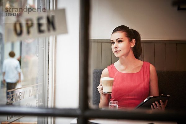 Junge Frau sitzend im Café mit digitalem Tablett und heißem Getränk