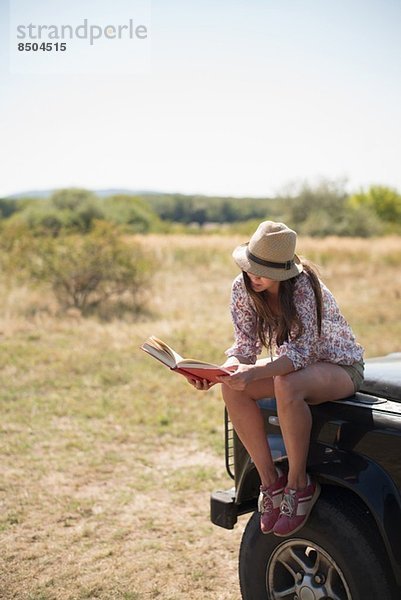 Mittlere erwachsene Frauen  die auf der Motorhaube des Autos sitzen und Buch lesen