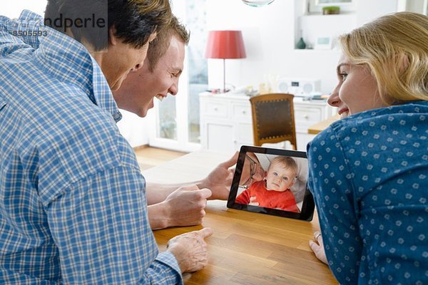 Großmutter und erwachsene Enkelkinder betrachten Foto des kleinen Jungen auf digitalem Tablett
