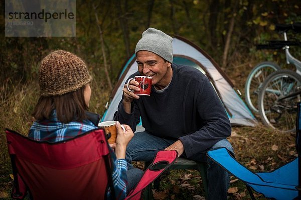 Reife Paare sitzen auf Campingstühlen vor dem Zelt und trinken heiße Getränke.