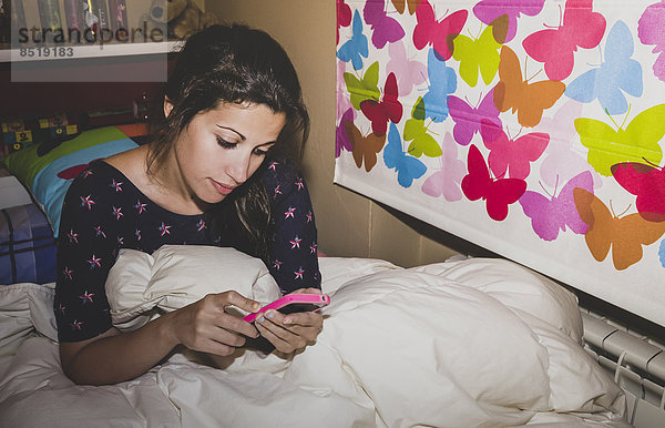 Spain  Madrid  young woman sitting on bed and using smart phone