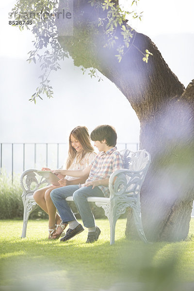 Bruder und Schwester mit digitalem Tablett auf der Bank unter dem Baum