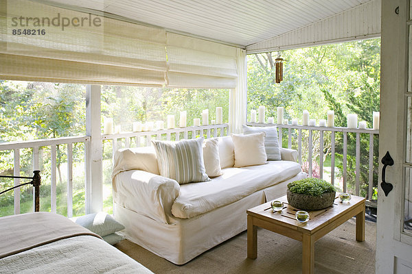 Eine Veranda oder schattige Veranda eines Hauses im Wald  mit Creme-Sofa und Kerzen entlang der Balustrade.