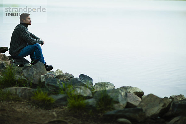 Ein Mann mittleren Alters sitzt auf Felsen und schaut nachdenklich über das Wasser.