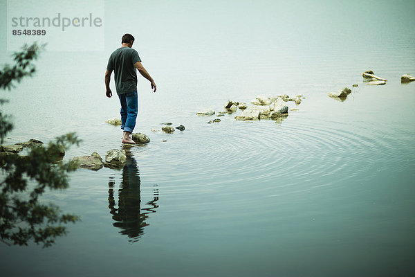 Ein Mann  der barfuß über Trittsteine vom Ufer eines Sees weggeht.