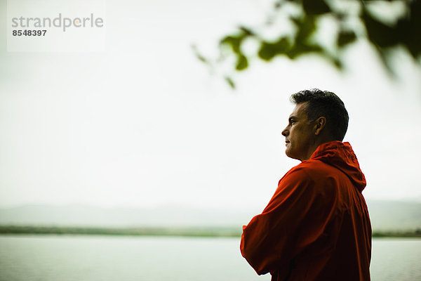 Ein Mann mittleren Alters in einer roten Jacke  der tief in Gedanken über das Wasser schaut.