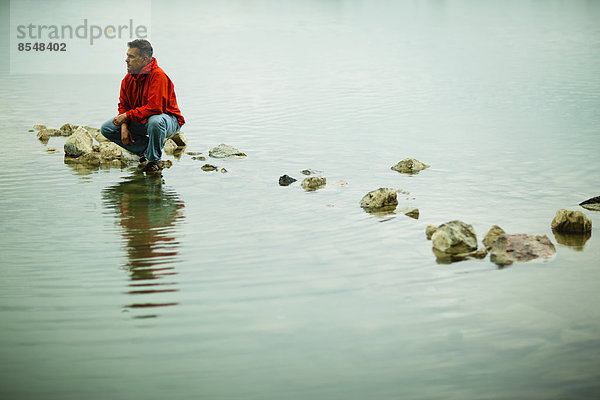 Ein Mann in einer roten Jacke balanciert in einer nachdenklichen Pose auf einem Trittstein oder Felsen im flachen Wasser.