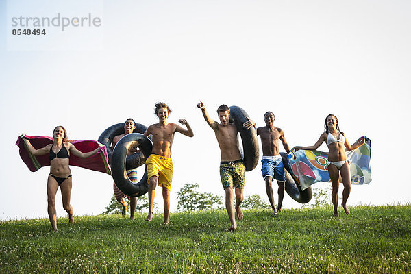 Eine Gruppe von Teenagern  Jungen und Mädchen  rannte mit Badetüchern und aufgeblasenen Schwimmern über das Gras.