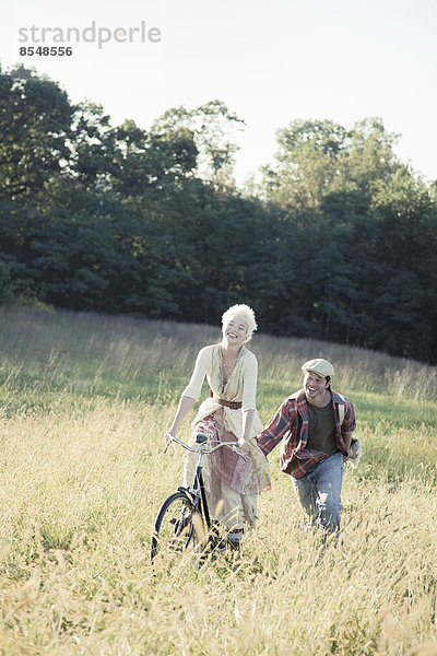 Ein Mädchen auf einem Fahrrad  das von einem jungen Mann durch das lange Gras geschoben wird.
