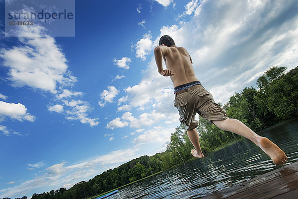 Ein Junge  der mit Anlauf von einem Holzsteg in ein ruhiges Wasserbecken springt.