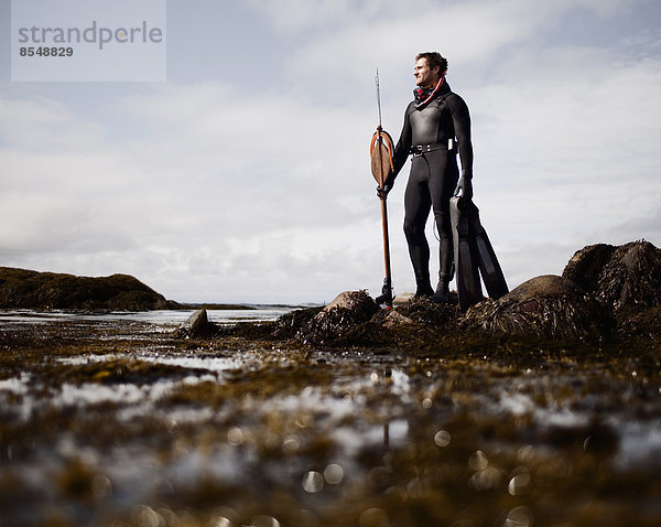 Ein Mann in einem Taucheranzug  der mit einer großen Speer-Fischerharpune am Ufer steht.