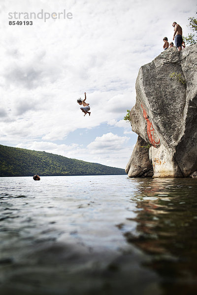 Eine Gruppe junger Leute springt von einer Klippe in das stille Wasser eines Sees.