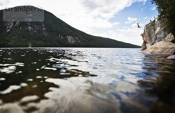 Eine Gruppe junger Leute springt von einer Klippe in das stille Wasser eines Sees.
