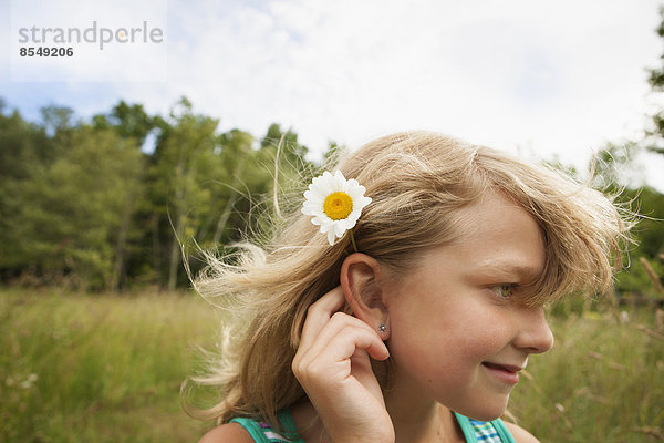 Ein junges Mädchen unter freiem Himmel  mit einer gänseblümchenähnlichen Blume hinter dem Ohr.