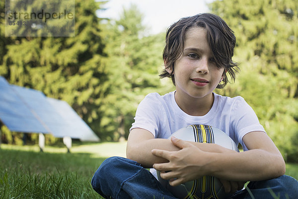 Ein Junge  der einen Fußball hält und auf dem Rasen sitzt. Sonnenkollektoren.