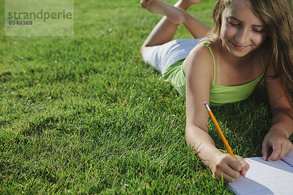 Lächelndes neunjähriges Mädchen sitzt im Gras und schreibt in ein Notizbuch