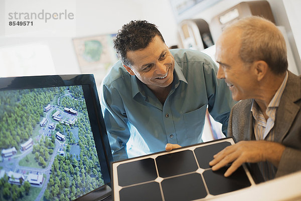 Zwei Männer in einem Architekturbüro. Sie arbeiten an einem grünen Bauprojekt und sitzen an einem Bildschirm. Ein Mann mit einer Fliesenprobe in der Hand.