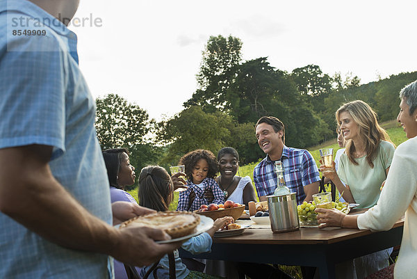 Eine Familie und Freunde  die an einem Tisch im Freien sitzen und eine Mahlzeit einnehmen.