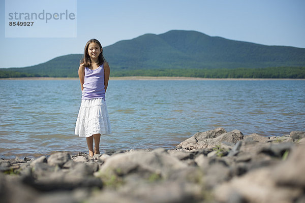 Ein junges Mädchen in weißem Rock und violettem Oberteil. Steht am Ufer eines Sees.