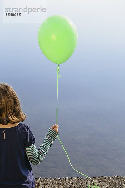 Neunjähriges Mädchen hält grünen Ballon in der Hand und sitzt am Wasser