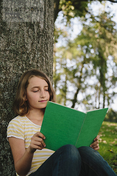 Neunjähriges Mädchen sitzt unter einem Baum und liest ein Buch